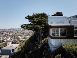 A modern residential building with concrete exterior, set on a hill overlooking a cityscape.
