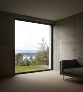 Modern, minimalist living room with a concrete wall and large window overlooking green hills and water.