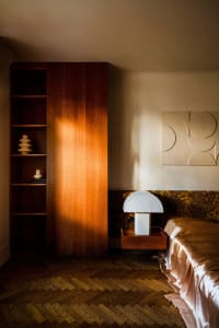 A minimalist bedroom with a wooden bookshelf, white lamp, and a bed with a fabric bedspread and headboard.