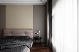 A modern bedroom with neutral-toned walls and furniture, featuring a bed with a dark gray duvet and pillows, a side table wit