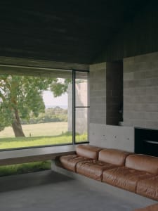 A modern living room featuring concrete walls and a leather sofa with a large window overlooking a grassy field.