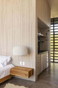 A minimalist bedroom with a wooden floating nightstand, white bedding, and a white pendant lamp hanging from a wooden wall pa