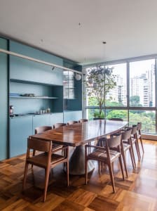 An industrial dining room featuring a wooden table and chairs against a blue wall with a tall glass window showing a cityscap