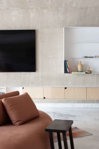 A minimalist living room featuring concrete-textured walls and a television unit with floating shelves and a bookcase below.
