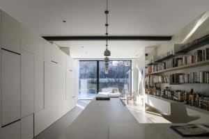 A minimalist home office features a white counter, tall cabinets, and a large, floor-to-ceiling window overlooking a garden.