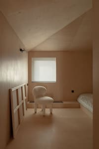 A minimalist bedroom with brown plywood walls, a window with blinds, and a small white chair.