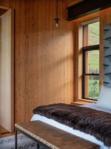 A minimalist bedroom with wooden walls, a fur blanket, and a window overlooking a green landscape.