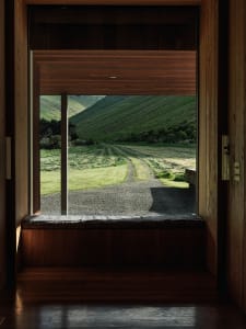 A view from the interior of a wooden building looking out through a window onto a rural road and hills.
