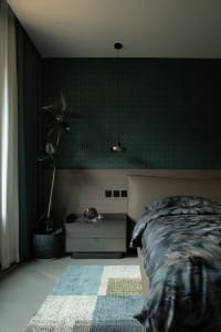 A modern bedroom featuring a bed with a mixed-material headboard, patterned bedding, and a side table with a single drawer. A