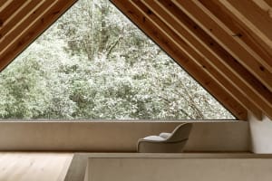 A minimalist wooden interior featuring a comfortable chair next to a large window overlooking a dense forest.