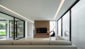 A modern living room with a large beige leather sofa, a wood-paneled wall with a television, and glass windows.