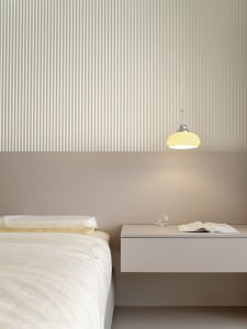 A modern minimalist bedroom featuring white bedding, a floating nightstand with a glass of water and an open book, and a pend