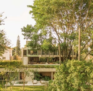 Modern residential house with glass exterior framed by lush greenery and trees.