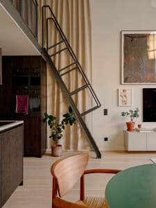 Modern loft interior with a steel ladder leading to a mezzanine level, featuring a kitchenette, wooden floor, and potted plan