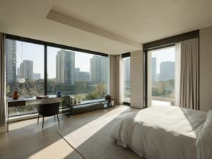 A minimalist bedroom with a large window overlooking city buildings and trees, featuring a bed with neutral linens and a simp