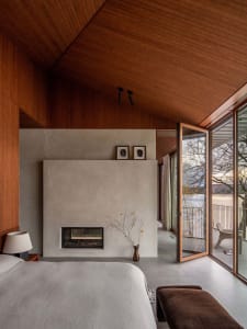 A modern bedroom with a concrete fireplace and large windows overlooking a serene outdoor view.