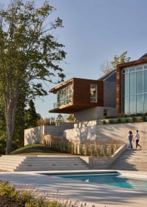 A modern residential building with stone and concrete exterior, wood accents, and a pool in front of it.