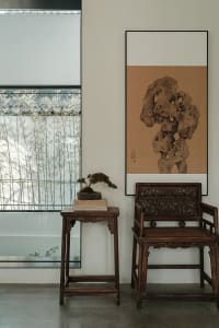 A modern living room featuring wooden furniture and stone accents, with a bonsai tree on a small wooden table and a framed ar