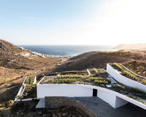 A modern, concrete residential building nestled in a rugged coastal landscape, featuring a rooftop garden overlooking the sea