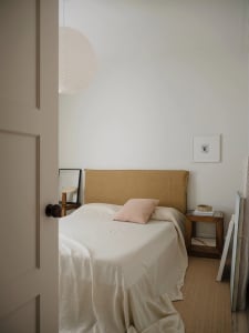 A minimalist bedroom with a bed covered in beige linens, a pink pillow, and a simple headboard.
