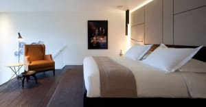 A minimalist bedroom featuring a large bed with a white cloth, an orange armchair, and a wooden floor with a gray rug.