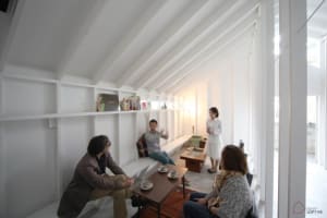 A minimalist living room featuring white walls, ceiling beams, and furniture, with people interacting in the space.
