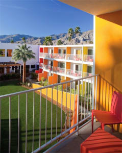 Hotel room balcony with metal railing, concrete floor, and glass wall overlooking palm trees and mountains.
