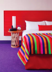 A modern bedroom featuring a vibrant, colorful striped blanket on a bed, with a red headboard and purple carpet.