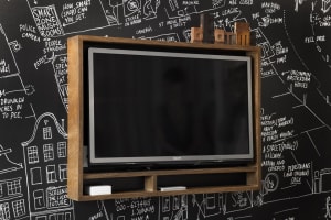 A wall-mounted electronics shelf with a wooden frame and a black flat-screen TV on it, against a chalkboard wall.