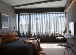 A minimalist bedroom featuring a textured bedspread, sheer curtains, and a large windowsill with a table and chair.