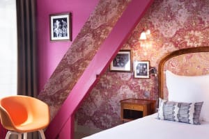 A cozy hotel bedroom with vibrant floral wallpaper, a wooden bed frame, and a modern orange accent chair.