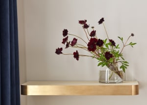 Glass vase with dark flowers on a brass shelf against a white wall.