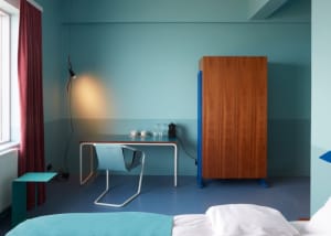 A minimalist bedroom with blue walls, wooden furniture, and a desk with a lamp and coffee mugs.