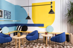 A modern cafe interior featuring a color-blocked wall with blue and yellow geometric shapes and blue cushioned seating.