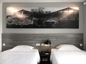 A modern hotel room features a wooden headboard with two twin beds, white linens, and a black lamp on a side table.