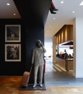A modern statue of a person dressed in a patterned suit and holding a suitcase stands in a lobby with black and white checker