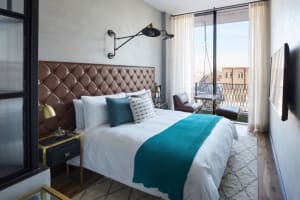 Modern minimalist bedroom featuring a bed with a tufted leather headboard, white bedding, and a teal throw blanket. The room 