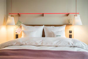 A modern, minimalist bedroom featuring a bed with white pillows and a pink throw blanket, a wooden headboard, and pendant lig