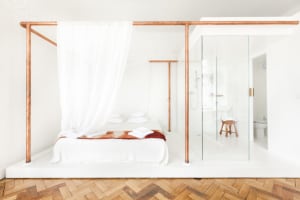 A minimalist bedroom featuring a white bed with wooden accents and a glass divider leading to a transparent bathroom