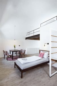 A minimalist bedroom with a single bed, wooden floor, metal ladder, and carpeted area rug.