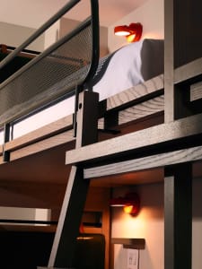 A minimalist, industrial-style bedroom featuring a wooden bunk bed with cabinets underneath and a modern red lamp.