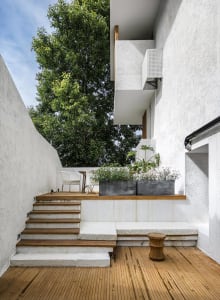 Minimalist outdoor space featuring wooden stairs, white benches, concrete walls, and metal planters with greenery.