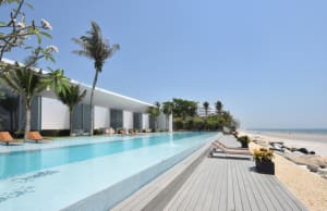 Modern glass and concrete beach resort with poolside palm trees and rock formations.