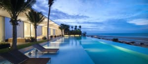 Modern residential house with glass walls and infinity pool by the beach at dusk.