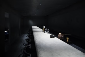 A minimalist restaurant bar with a concrete countertop and black seating on a dark tiled floor.