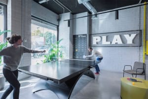 A modern playroom featuring concrete walls and a ping pong table with a 'PLAY' sign and a 'Hall of Fame' scoreboard.