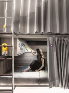 Person sitting on a minimalist bunk bed with a wooden frame and curtains.