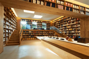 A spacious library with multiple shelves filled with books, illuminated by ceiling lights and natural light through large win