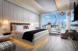 Modern hotel room with bed, desk, chairs, and large windows overlooking a cityscape.