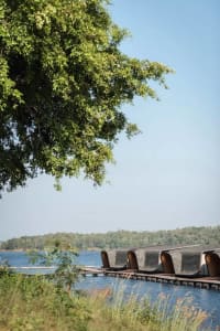 A series of wooden floating huts on a calm lake surrounded by lush greenery under a clear blue sky.